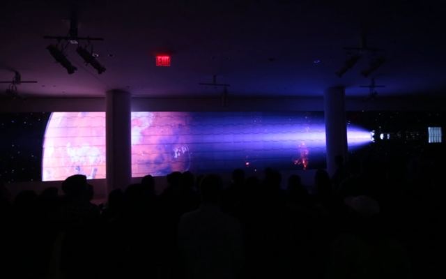 Video still: a dark hall lit by a wide projected planet