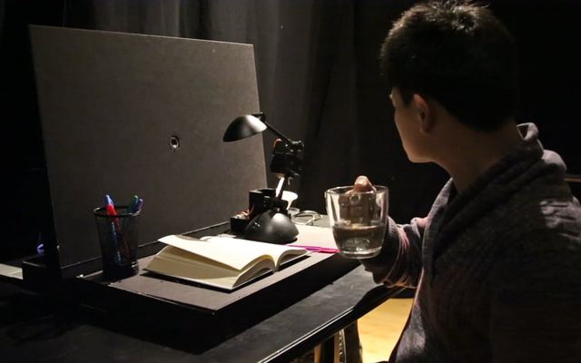 Video still: a person at a desk with a lamp, a book and a glass